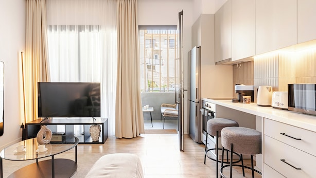 Una sala de estar moderna con un televisor de pantalla plana, una mesa de centro de vidrio y un sofá. Se observa una zona de cocina con un refrigerador, estufa y taburetes de bar.