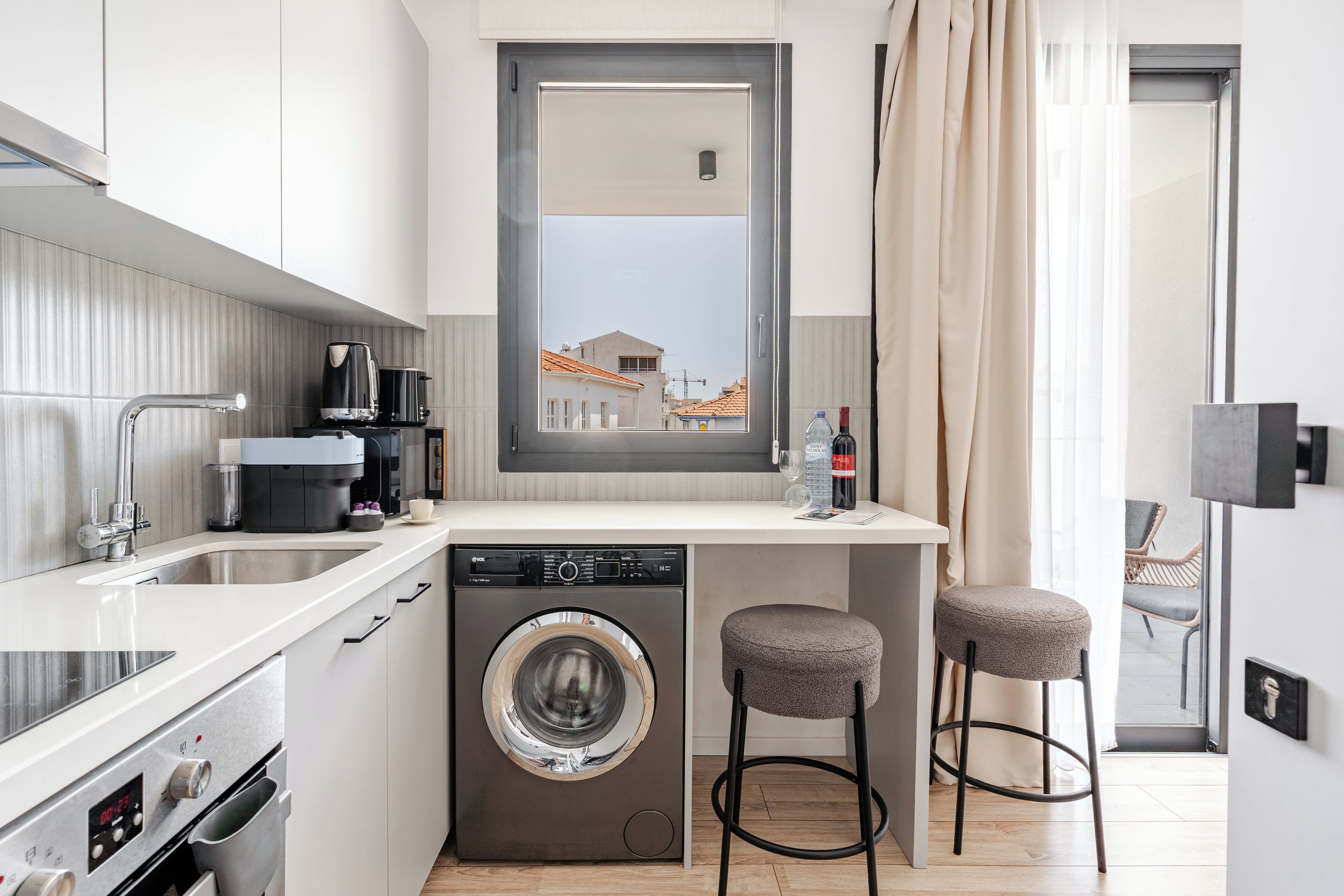 Een compacte keuken met een wasmachine, een koffiezetapparaat en een klein barretje met krukjes.
