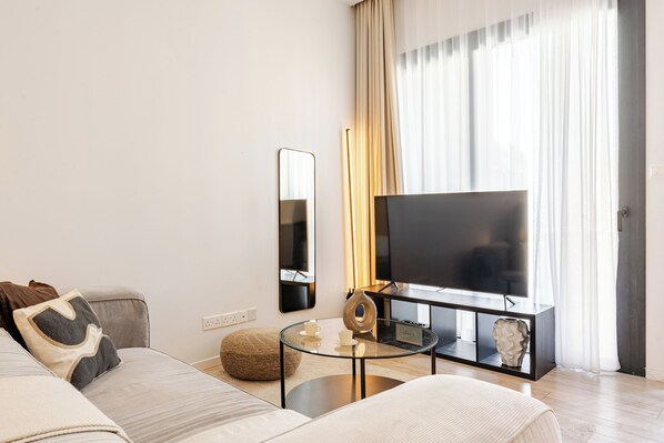 Una sala de estar moderna con un sofá, una mesa de centro de vidrio, un televisor de pantalla plana y una mesita auxiliar con objetos decorativos.