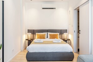 A modern hotel room with a large bed, two bedside tables with lamps, an air conditioning unit, and a wooden floor.