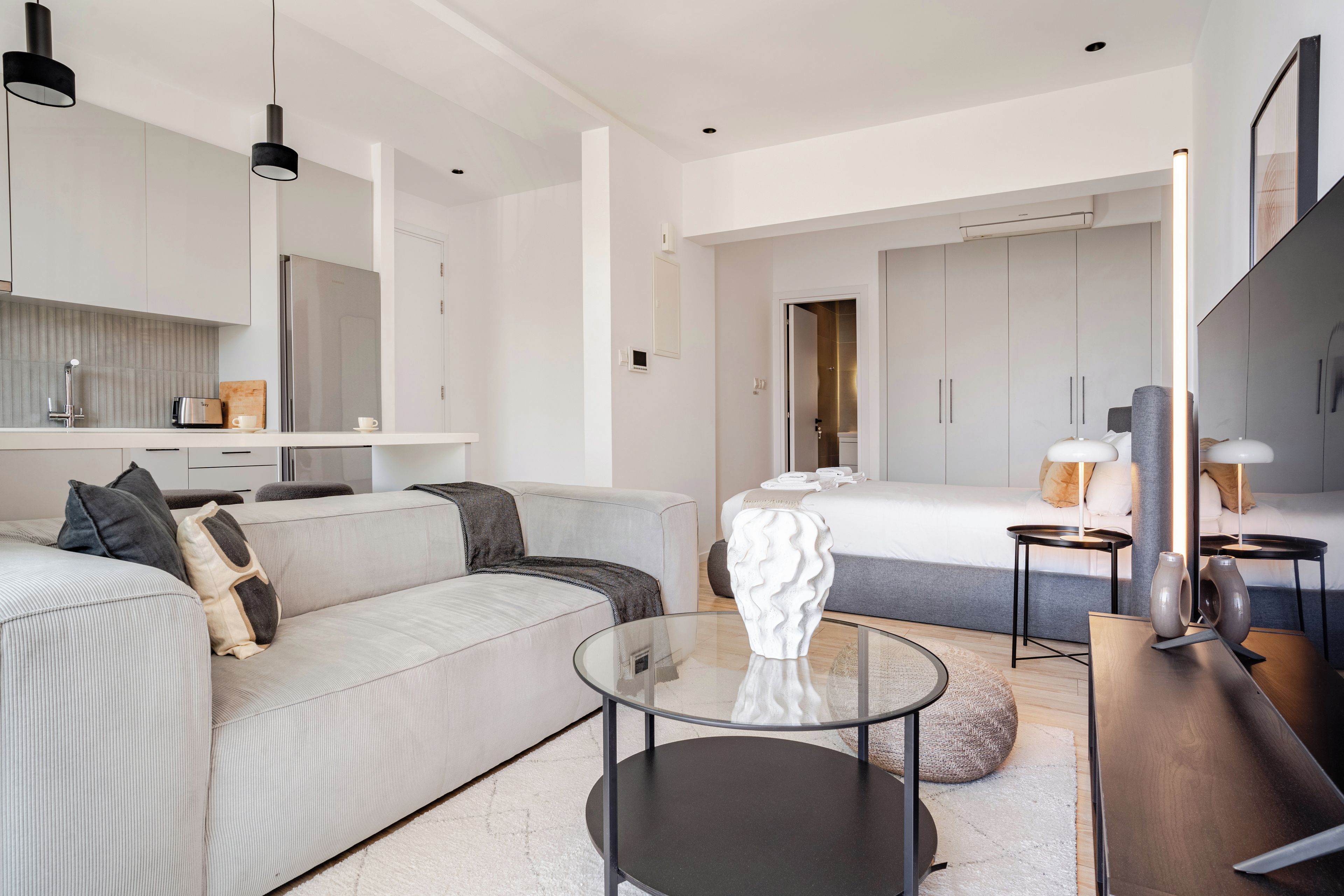 Una habitación de hotel moderna con sofá, una mesa de centro de vidrio, una cama con cabecero, una mesita de noche con lámparas y un espejo grande.