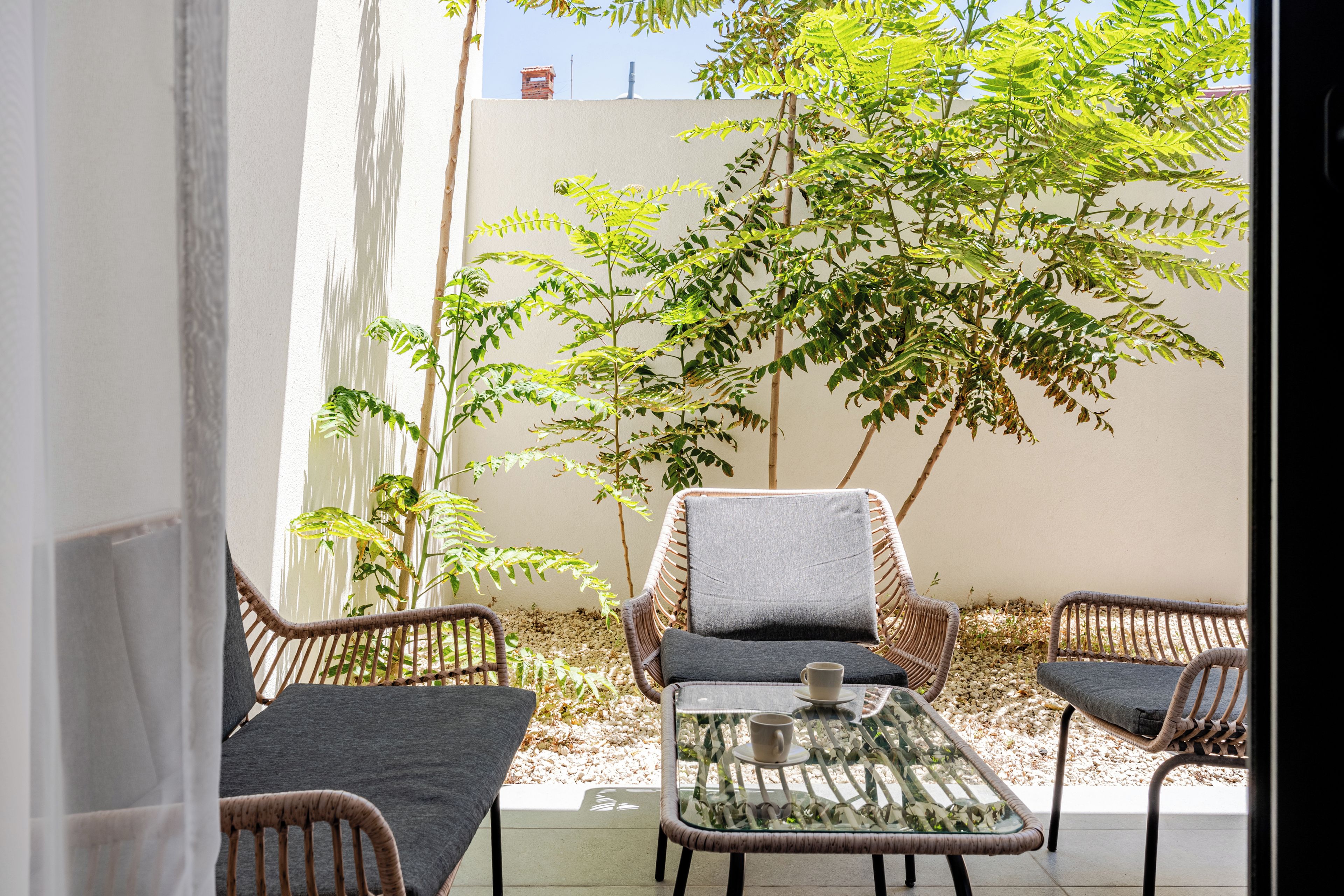 An outdoor seating area with wicker chairs and a glass table, surrounded by greenery and a white wall.