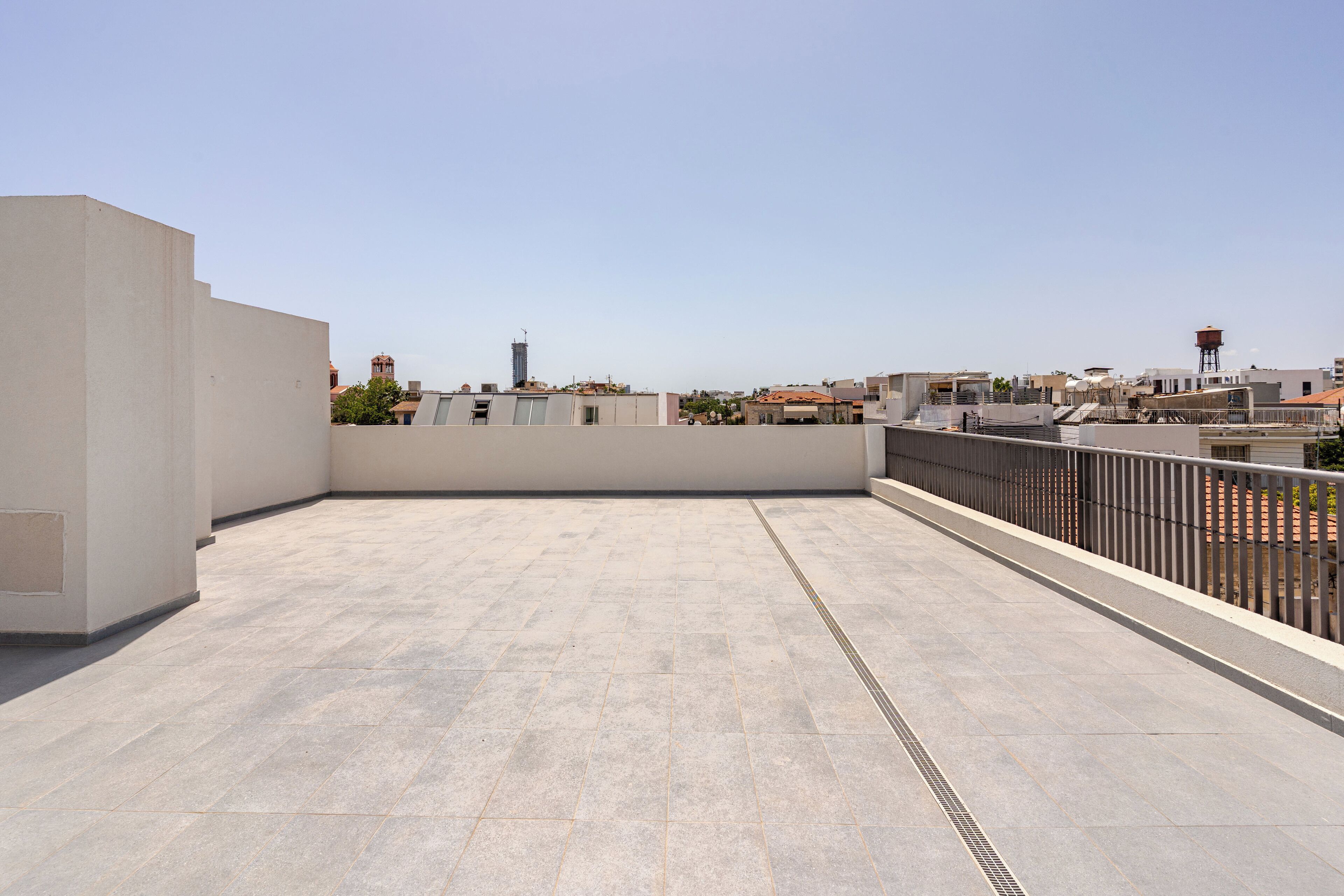 A rooftop terrace with a tiled floor, a metal railing, and a view of surrounding buildings.