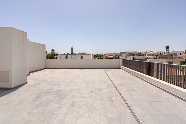 Una terraza en la azotea con suelo de baldosas, barandilla metálica y vistas de edificios circundantes.