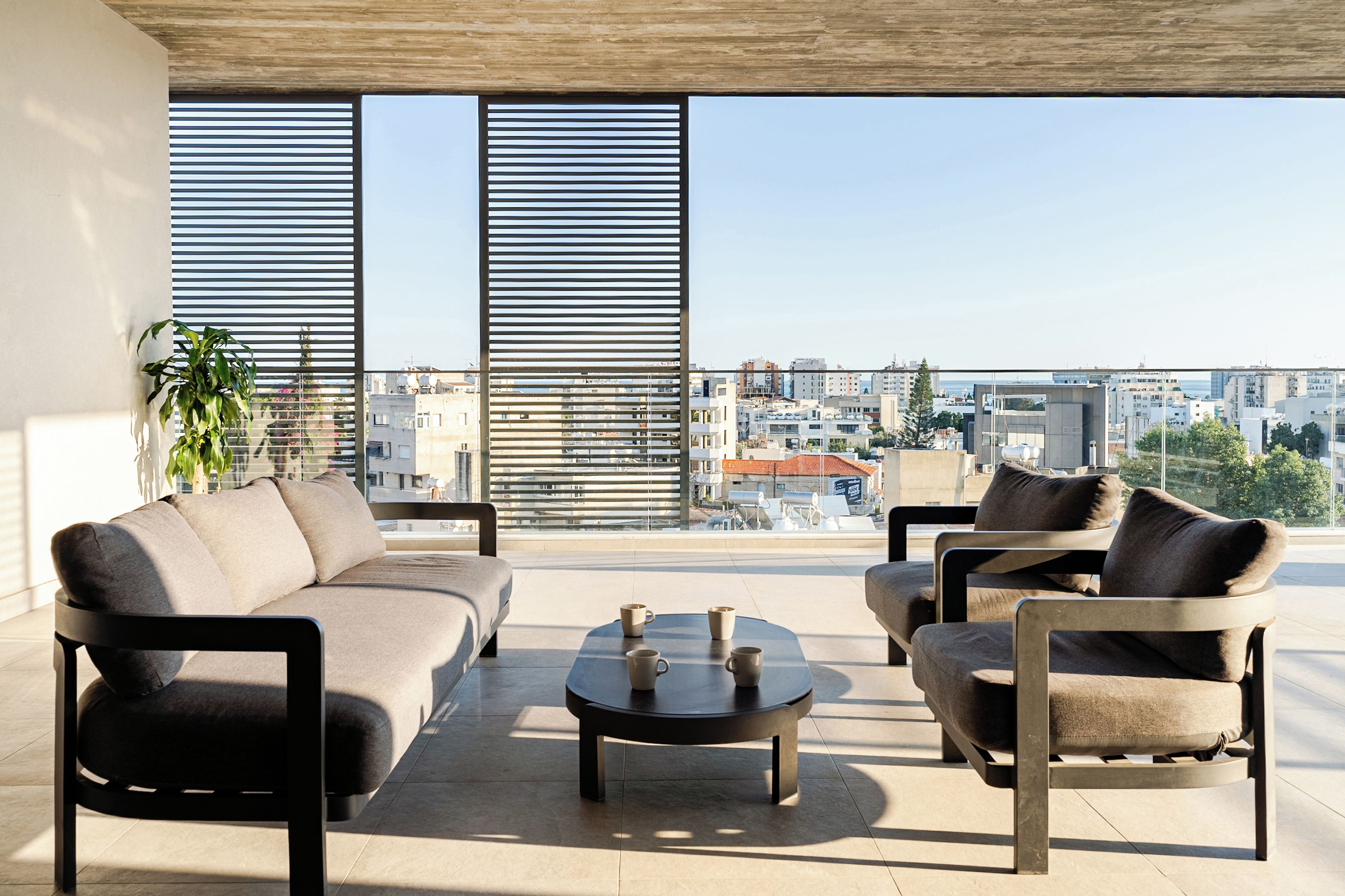 Una sala de estar moderna con sofá, sillones y una mesa de centro, con ventanas grandes con persianas horizontales y vistas a la ciudad.