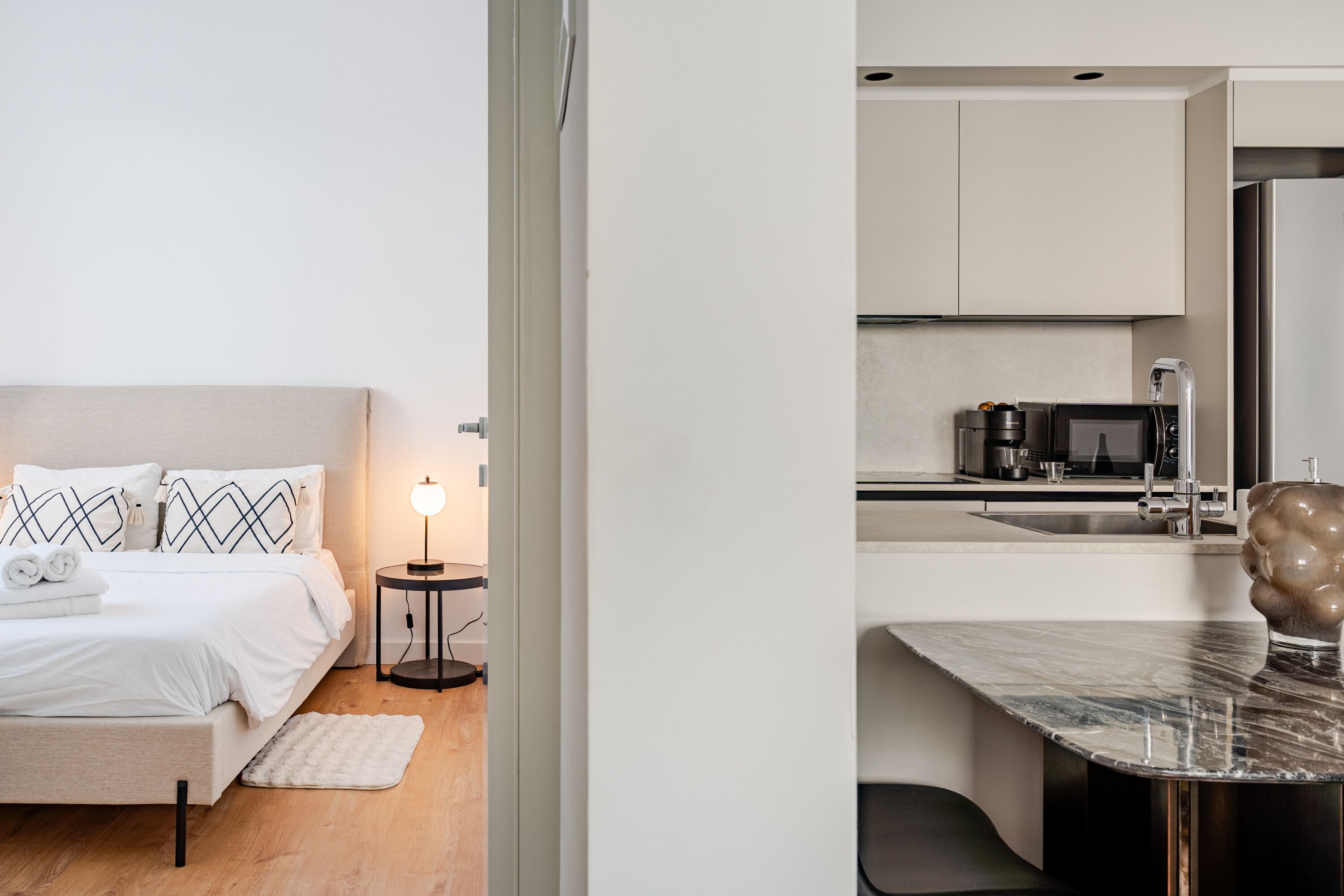 A modern hotel room with a bed, bedside table, lamp, and a view into the kitchen area.