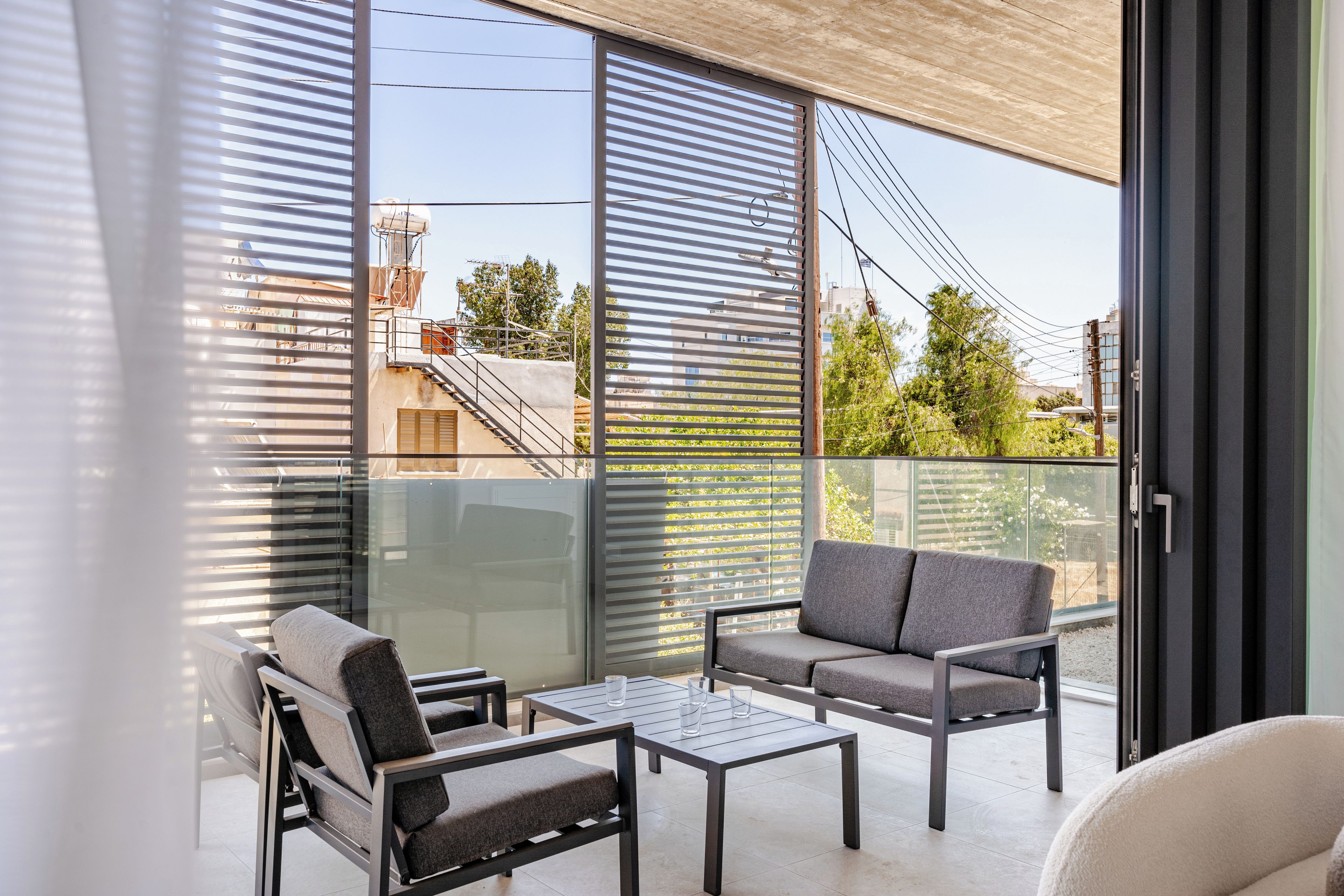 Ein Balkon mit einer Couch, zwei Stühlen und einem Tisch, mit großen Fenstern und Blick auf Gebäude und Bäume.