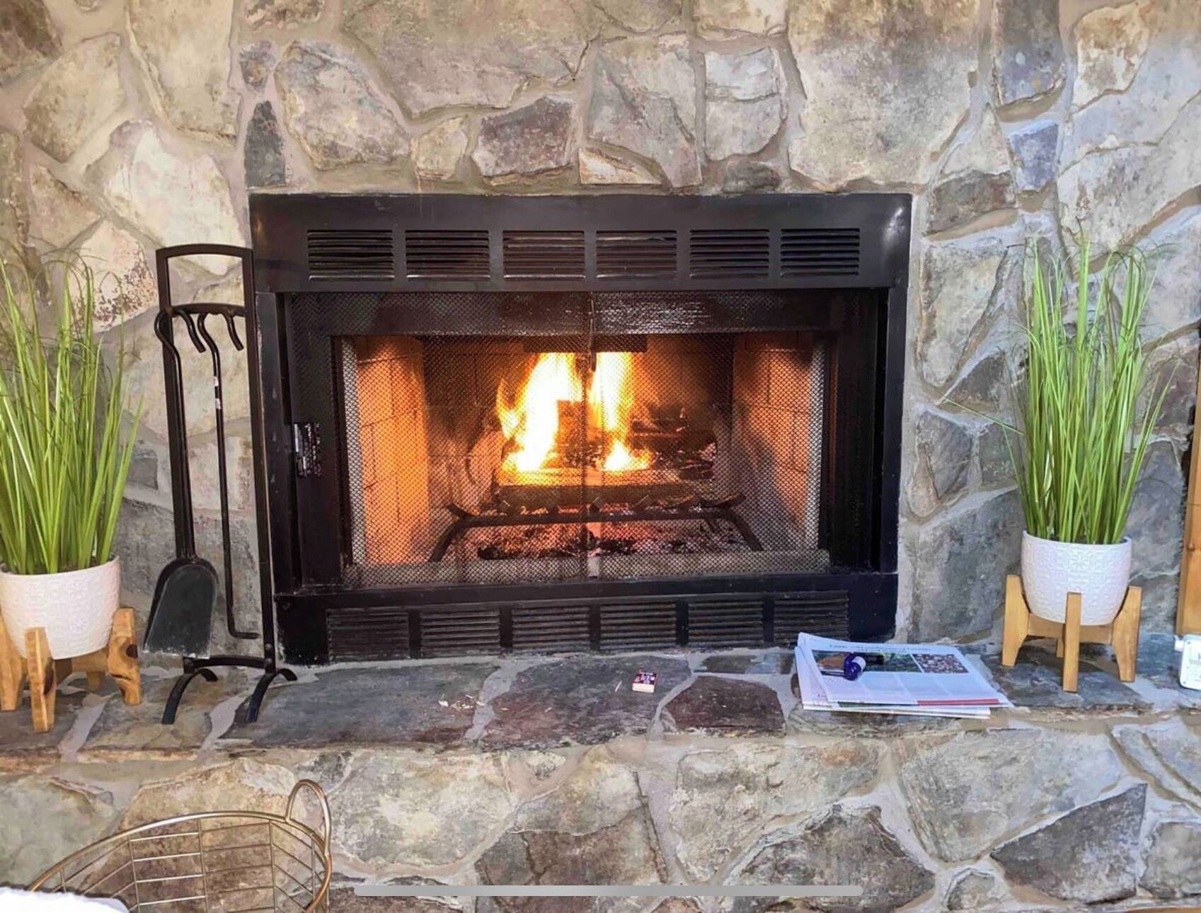 A stone fireplace with a fire, flanked by potted plants and a chair.