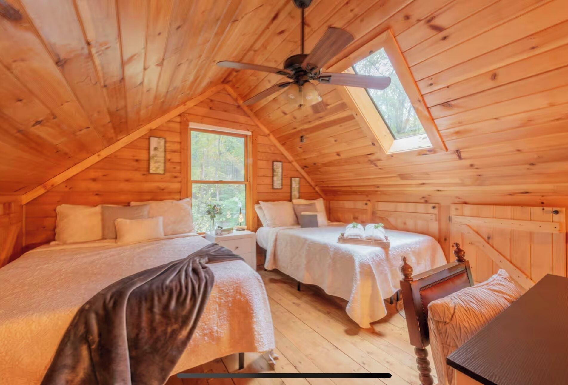 A cozy attic bedroom with two beds, a ceiling fan, and large windows.