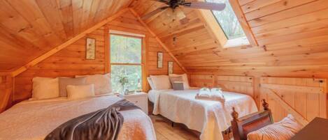 A cozy attic bedroom with two beds, a ceiling fan, and large windows.