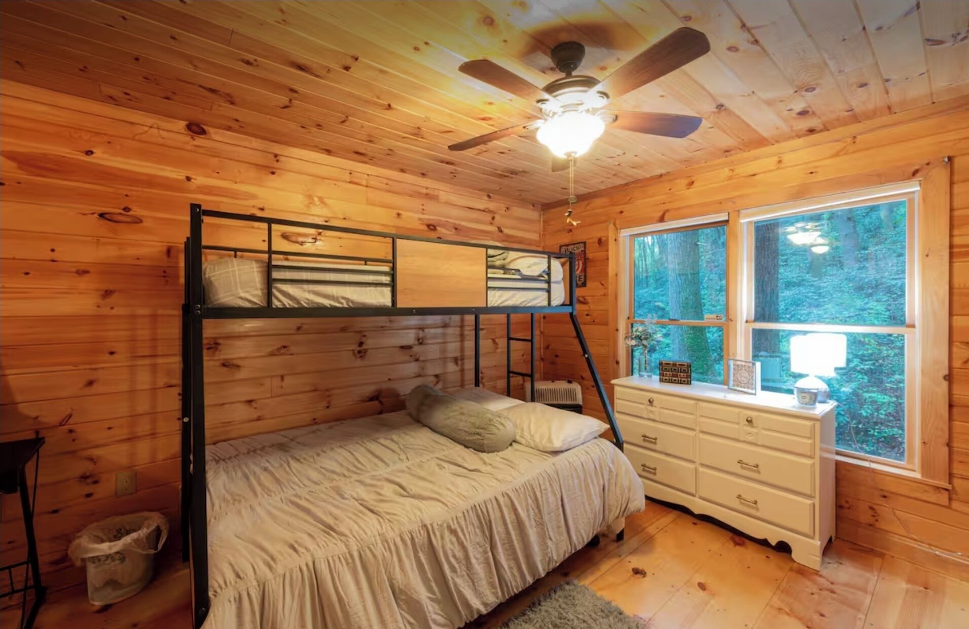 Un dormitorio de cabaña de madera con una cama litera, un aparador y una ventana con vistas al bosque.