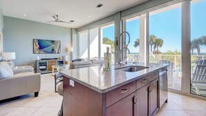 Een moderne keuken met een groot eiland, een zithoek met een bank, en een uitzicht op palmbomen door grote ramen.