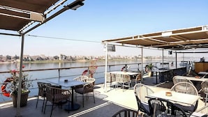 Terraza exterior con mesas y sillas, con vistas a un cuerpo de agua y un paisaje urbano.