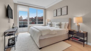 A modern bedroom with a large bed, a bedside table with a lamp, a TV mounted on the wall, and a view of the city through large windows.