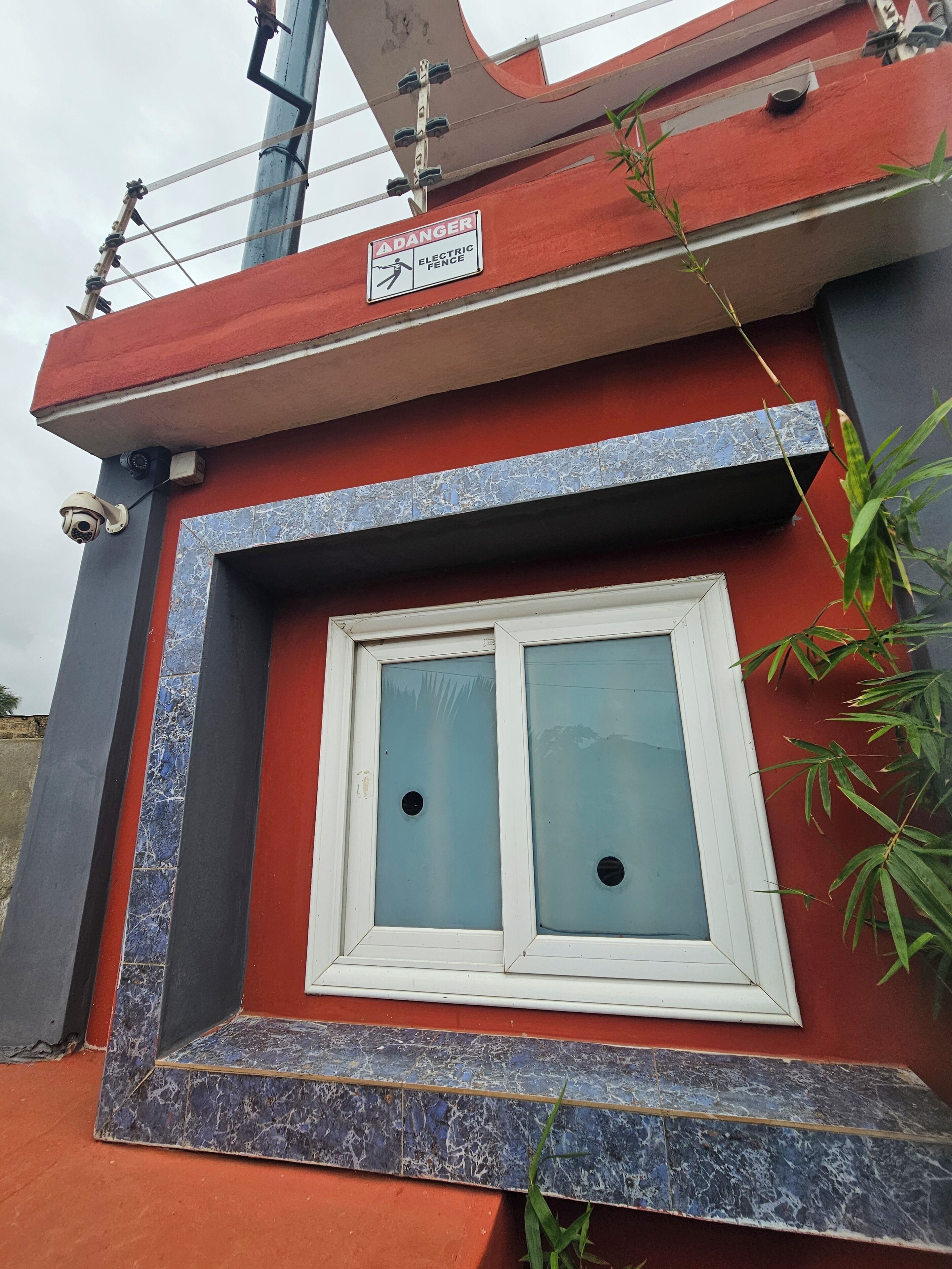A building with a red and grey facade, a window with two panes, and a 'DANGER ELECTRIC FENCE' sign.