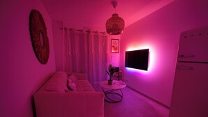 Een kamer in roze verlichte sfeer, met een bank, een tafeltje met een plant, een tv aan de muur en een koelkast.