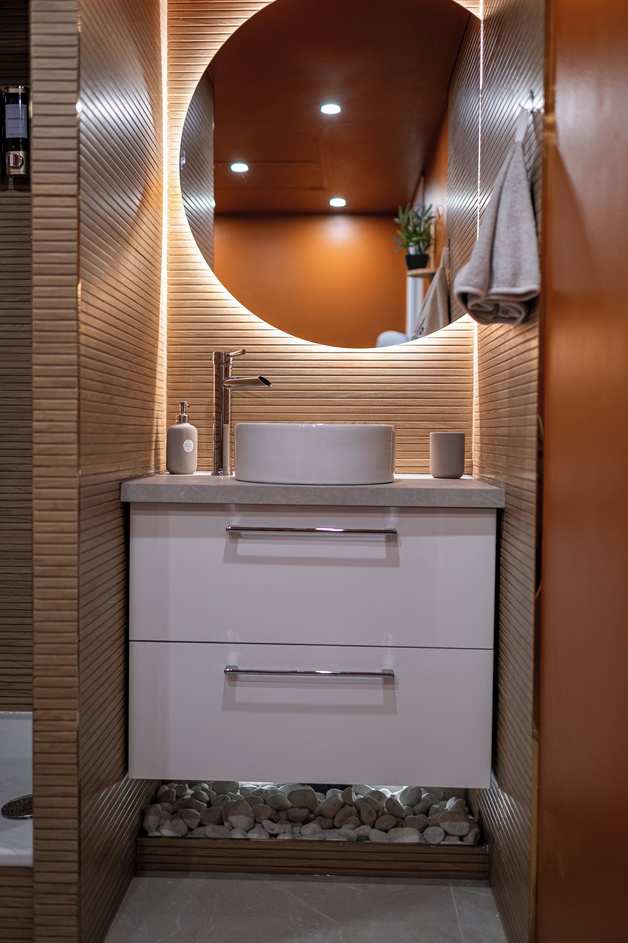 Een moderne badkamer met een ronde spiegel, een witte wastafel en een wandpaneel van hout.