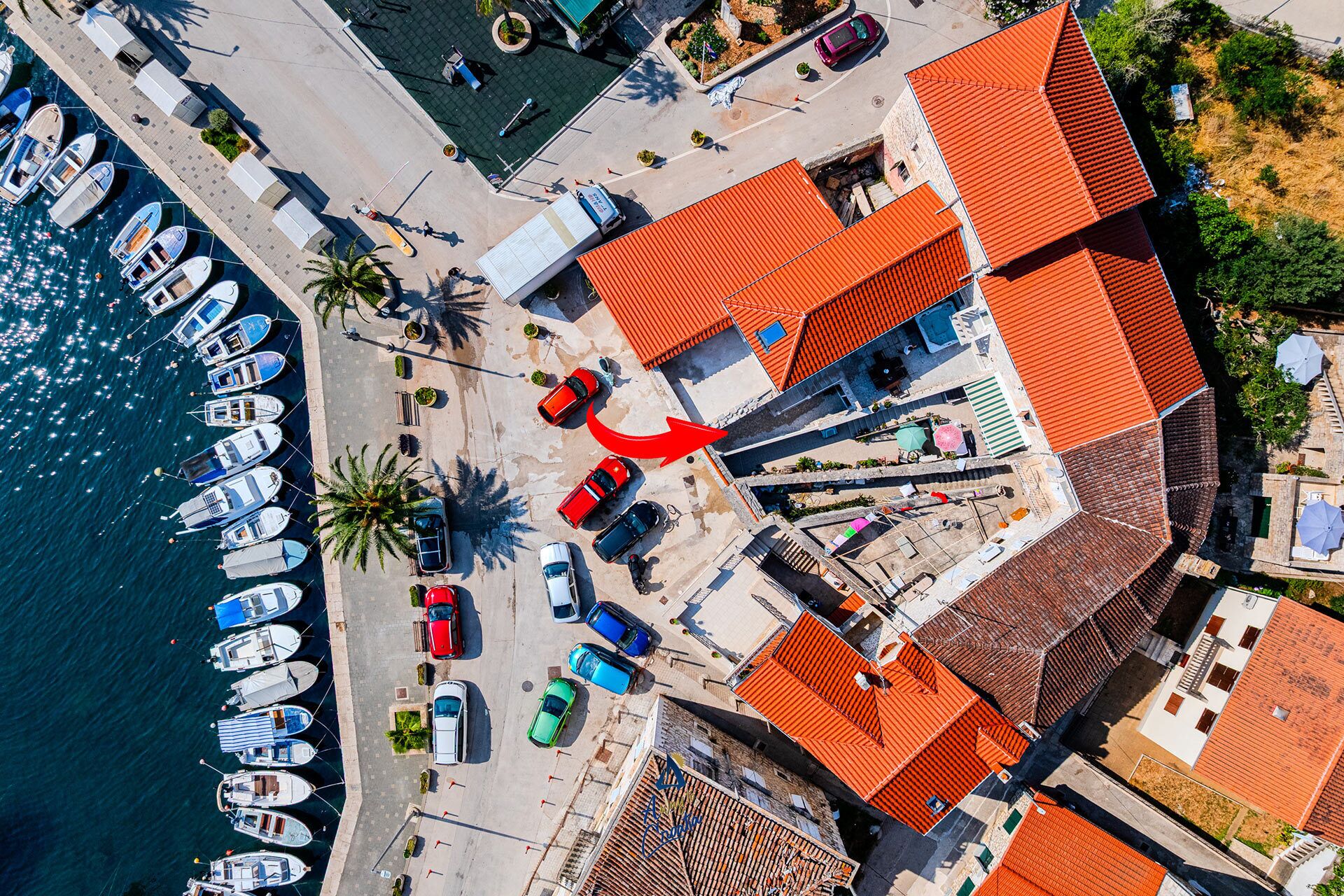 An aerial view of a coastal area with boats docked along the waterfront, a red car on the road, and buildings with red roofs.
