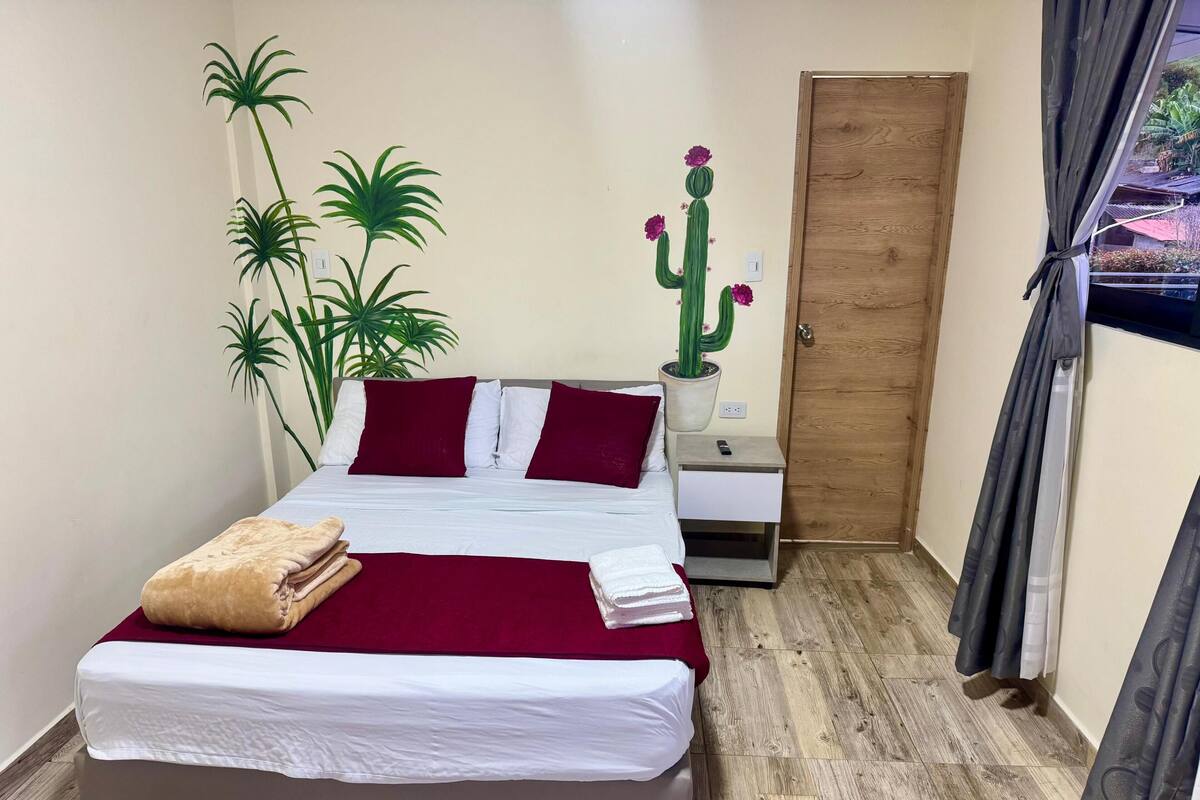 A neatly made bed with a red and white bedspread, a wooden nightstand with a remote control, a potted cactus, and a door to the right.