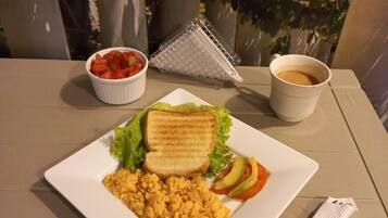A breakfast plate with scrambled eggs, toast, lettuce, tomato, and avocado, a cup of coffee, and a grater on a table with a napkin.