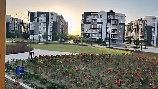 Ein Wohngebiet mit modernen Mehrfamilienhäusern, einem grünen Park und einem kleinen Garten mit roten Blüten.