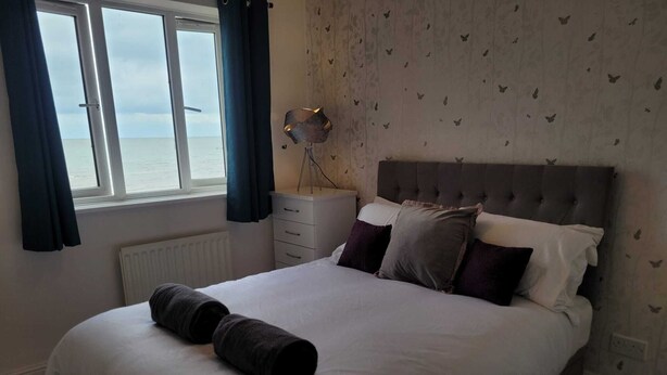 Une chambre à coucher avec un grand lit, une table de chevet, une lampe et une vue sur la mer par la fenêtre.
