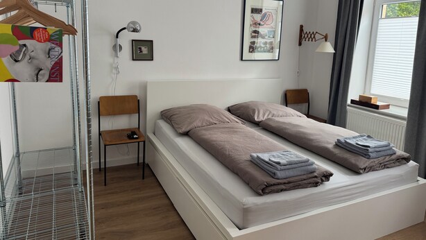 Un dormitorio moderno con una cama blanca, dos almohadas, una mesita de noche, una silla, un perchero y una ventana con cortinas.
