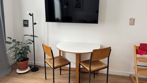 Una habitación moderna con una televisión de pantalla plana en la pared, una mesa redonda blanca, dos sillas de madera, una planta en maceta y una lámpara de pie.
