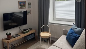 Una sala de estar con una televisión de pantalla plana, una mesita de madera, una silla blanca, un sofá beige con un cojín azul y una ventana con cortinas.