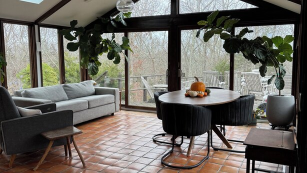 A spacious room with a high ceiling, a large window, a dining table with chairs, a sofa, and a small side table.