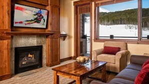 A cozy living room with a fireplace, a television mounted on the wall, and a large window offering a view of a snowy landscape.