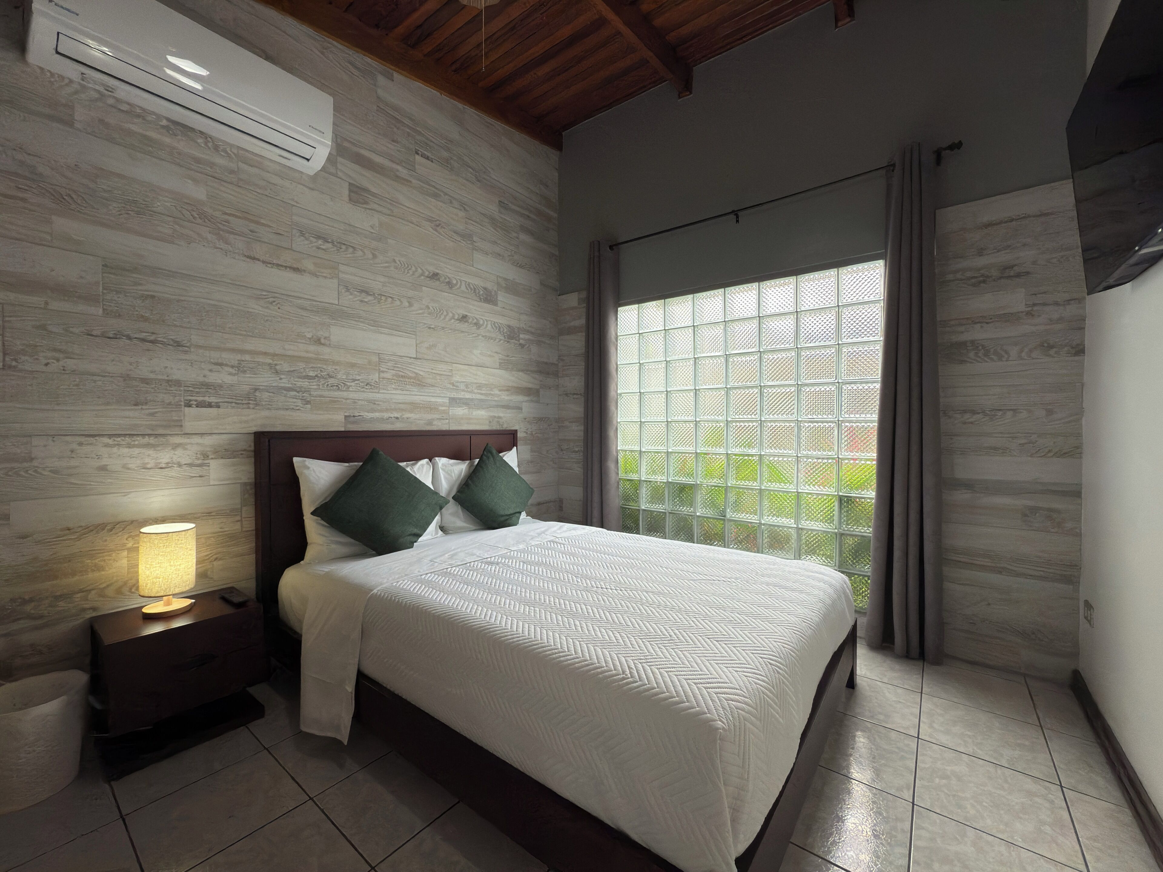 A bedroom with a wooden headboard, a bed with white bedding, a nightstand with a lamp, and a large window with a view.