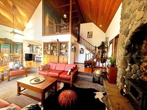 A spacious living room with a high ceiling, a fireplace, a staircase, and a large bookshelf.
