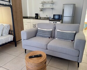 A modern living room with a grey sofa, striped pillows, a remote control on a woven stool, a kitchen area with a microwave, coffee maker, and a refrigerator.