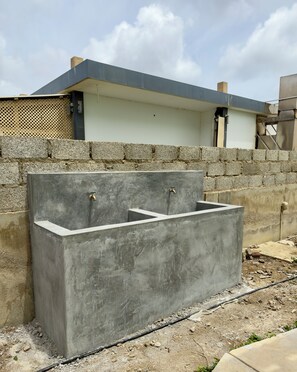 A concrete water feature with two taps, set against a stone wall and a building with a flat roof.