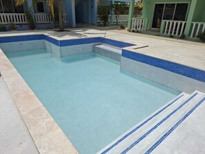 A rectangular swimming pool with blue-tiled edges, surrounded by a concrete deck and a white fence.