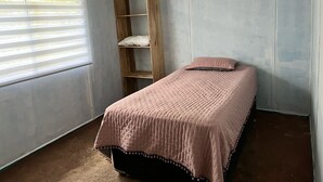 Ein kleines Zimmer mit einem Einzelbett, einem Bücherregal aus Holz, einem Fenster mit Jalousien und einer rosa Bettdecke.