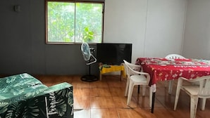 Ein Zimmer mit Fliesenboden, einem Fernseher auf einem Schrank, einem Ventilator, einem Tisch mit roter Tischdecke und einem Fenster mit Blick ins Grüne.