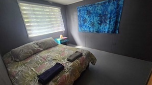 Ein Schlafzimmer mit einem Bett, einem Nachttisch, einem Fenster mit Jalousien und einem blauen Wandteppich.