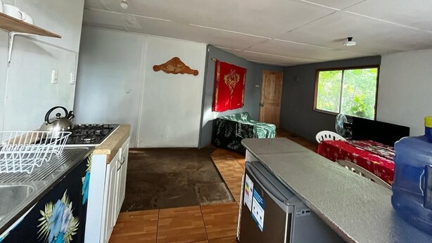 Eine kompakte Küche mit Gasherd, Wasserkocher und kleinem Kühlschrank. Ein Essbereich mit Tisch und Stühlen ist sichtbar, und ein Fenster sorgt für natürliches Licht.
