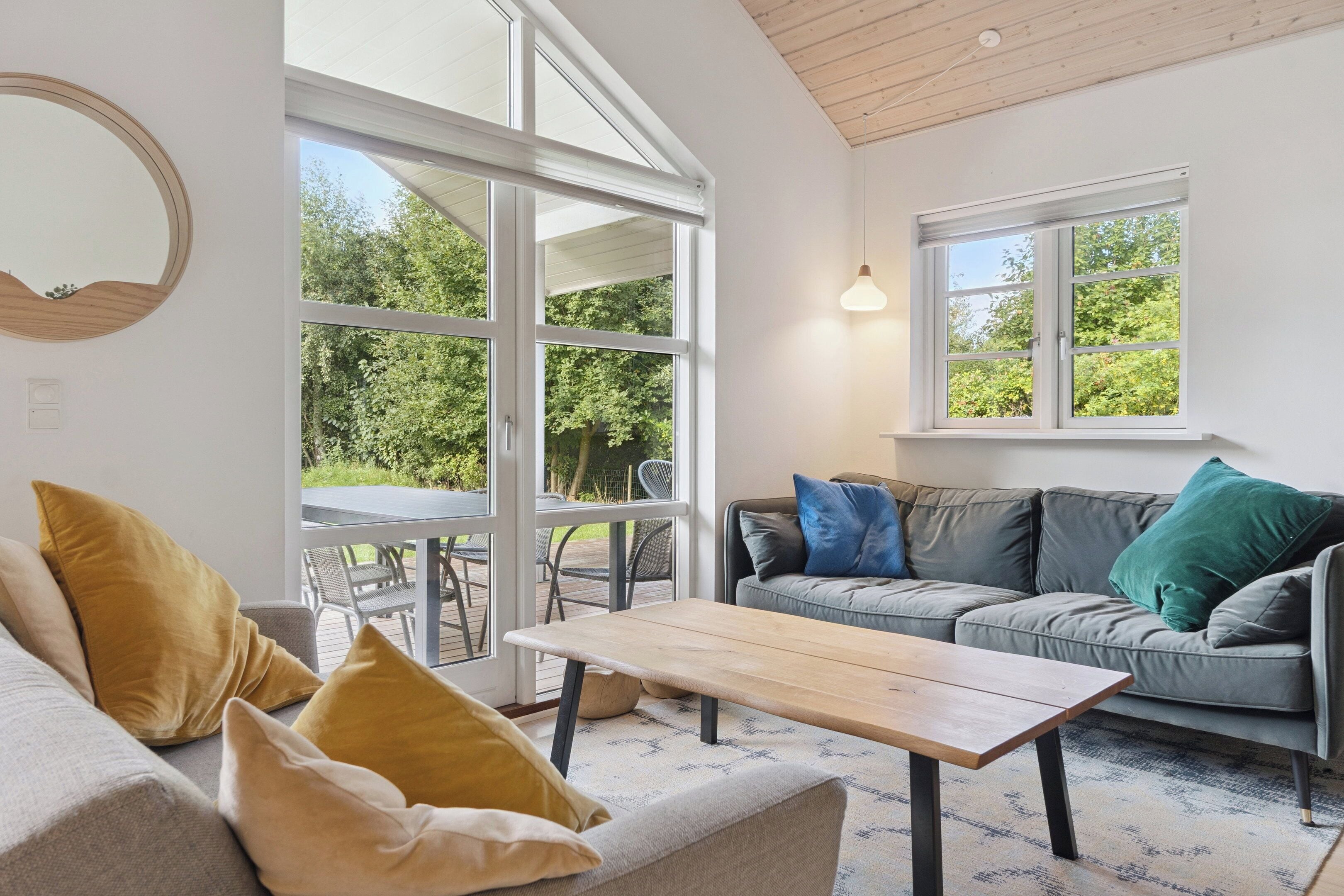 En moderne stue med sofa, sofabord og store vinduer med udsigt til grønne områder.