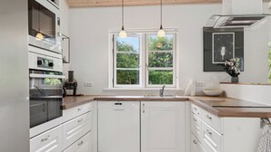 Une cuisine moderne avec des meubles blancs, un four encastré, un lave-vaisselle et une fenêtre donnant sur un espace verdoyant.