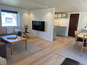 A modern living room with a sofa, a coffee table, a TV mounted on the wall, and a kitchen area with cabinets and a sink.