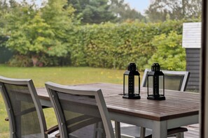 A wooden table with chairs, two lanterns, and a view of a garden with a hedge and trees.