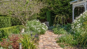 A garden pathway with a brick walkway, surrounded by various plants and a white gazebo.