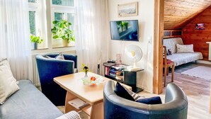 A modern living room with a sofa, armchair, and a dining table. There is a TV mounted on the wall, a fan, and a window with curtains. - Zum Storchennest by Interhome (Chemnitz)