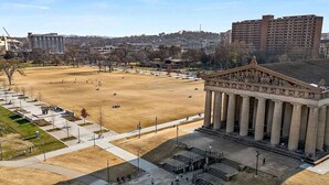 An open public square with a classical building featuring columns, surrounded by a few people and modern buildings in the background.