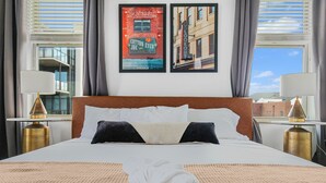 A hotel room with a large bed, two nightstands, a window with blinds, and two framed pictures on the wall.
