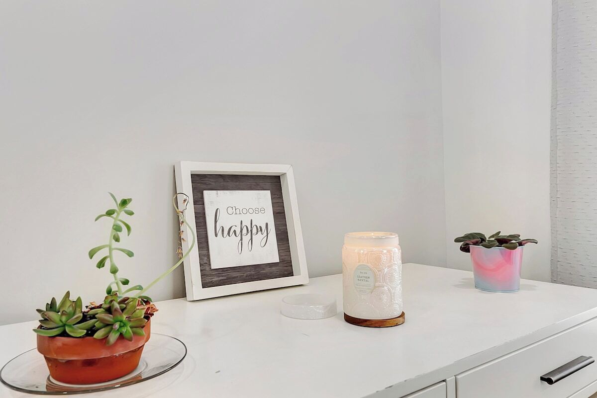Uma mesa branca com uma placa emoldurada, uma vela e duas plantas em vasos.