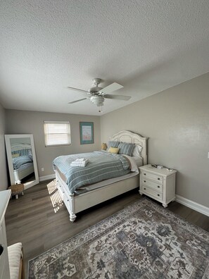 Une chambre à coucher comprenant un lit, des tables de chevet, une commode, un ventilateur de plafond, une fenêtre avec des stores et un tapis.